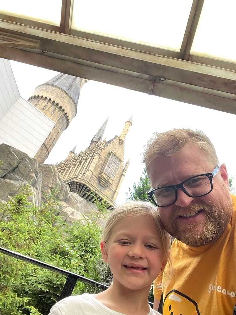 A man with glasses and a beard and a young blonde girl smile at the camera in a selfie, with the stone towers of Hogwarts Castle visible behind them through a window frame, surrounded by green trees.