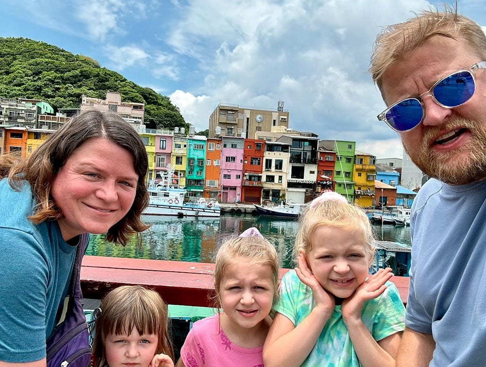 A family selfie on a boat at Zhengbin fishing harbor in Keelung, Taiwan, with a row of brightly colored stacked buildings and moored boats visible across the water behind them.