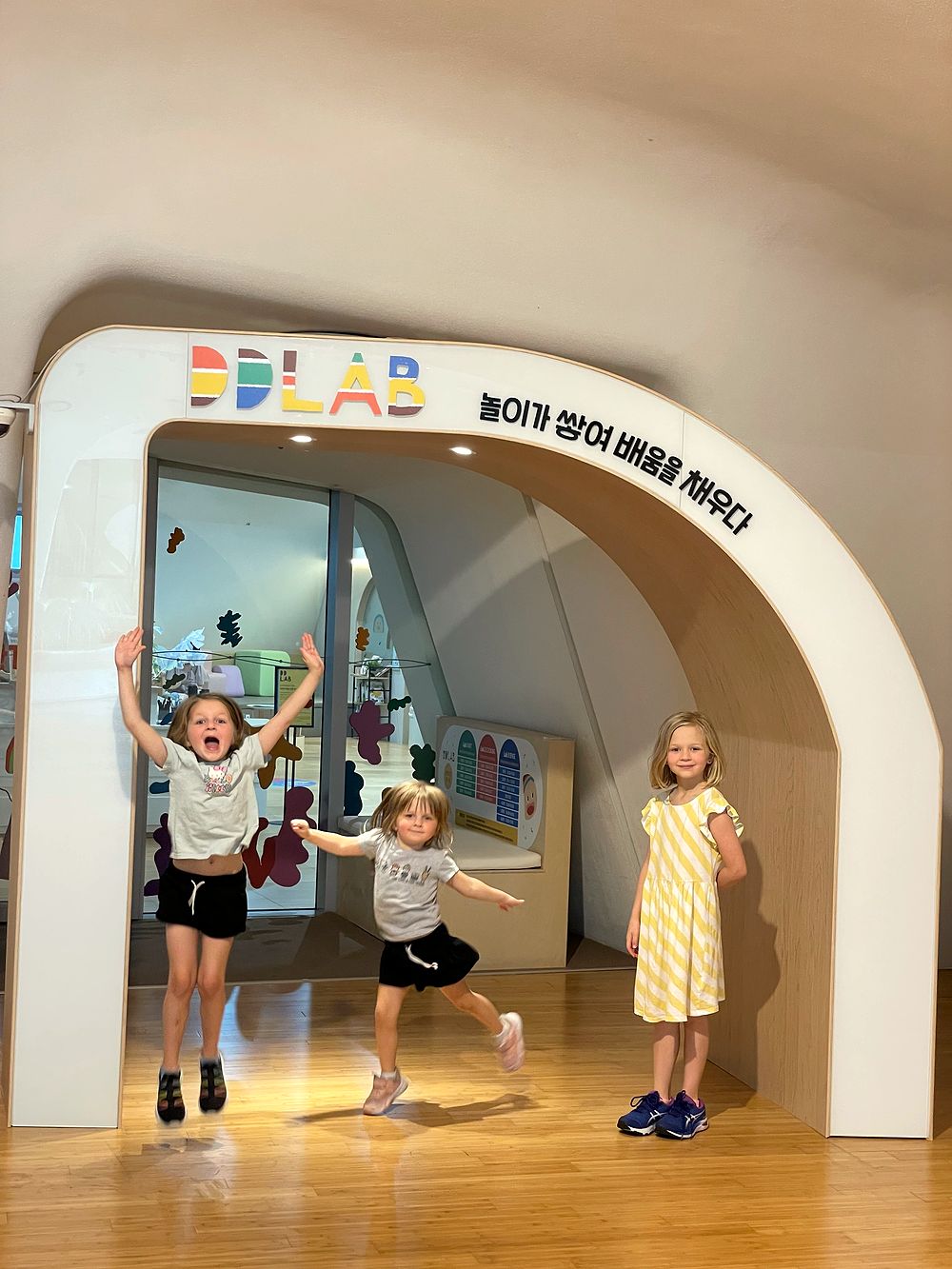 Three girls at the rounded archway entrance of DDLAB children's museum in Seoul, two of them mid-jump with arms raised
