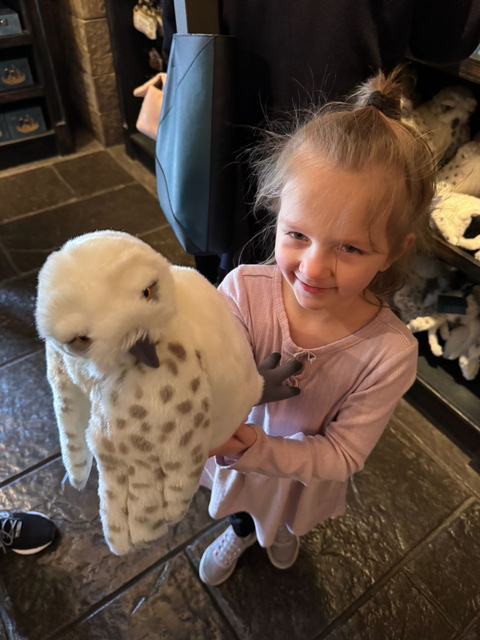 A young girl in a pink dress smiles while holding a large white snowy owl stuffed animal inside a Harry Potter World merchandise shop.