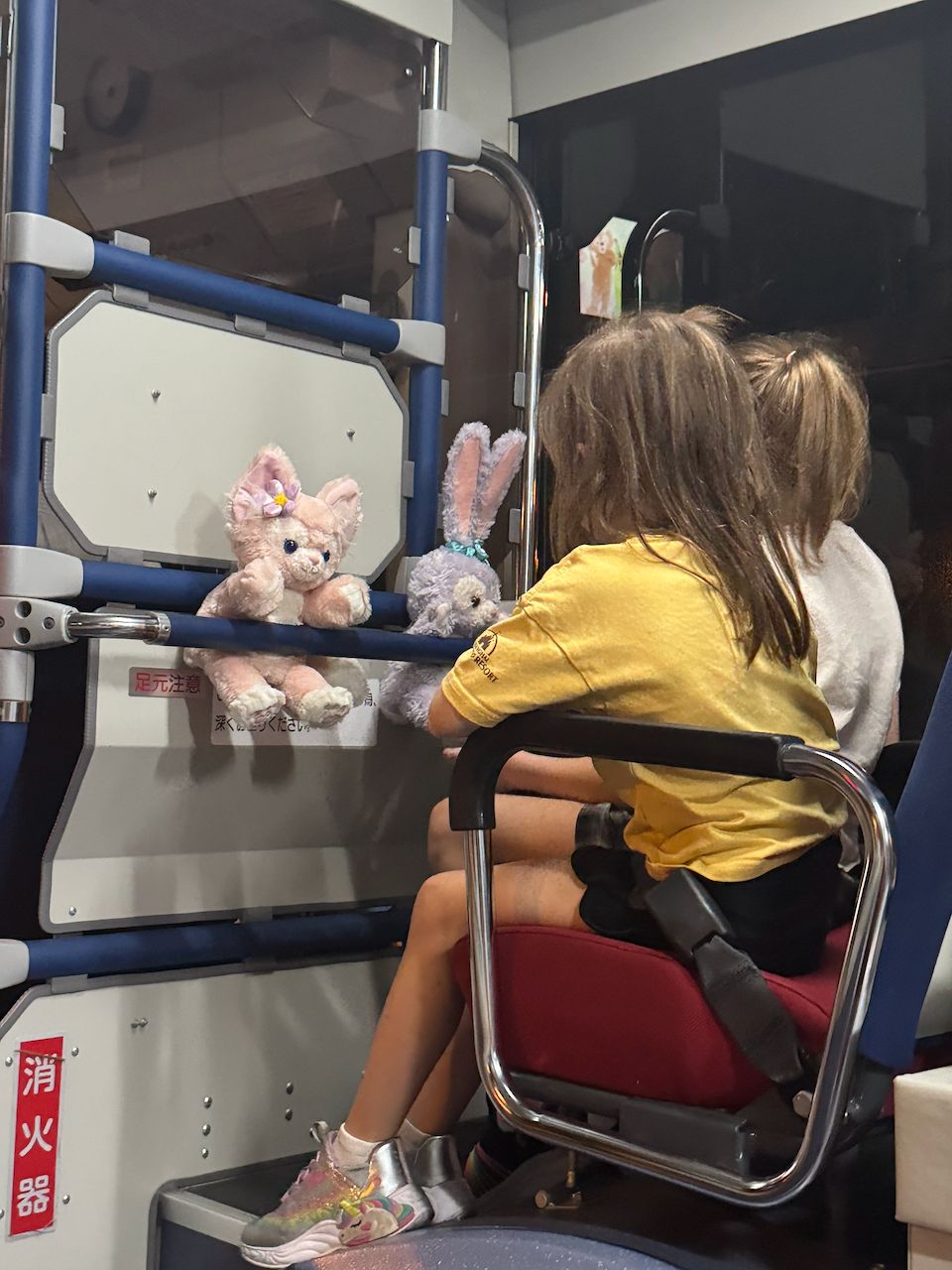 A young girl in a yellow shirt sits on a monorail seat with two Disney plush toys propped against the railing in front of her, Japanese safety signage visible on the wall beside them.