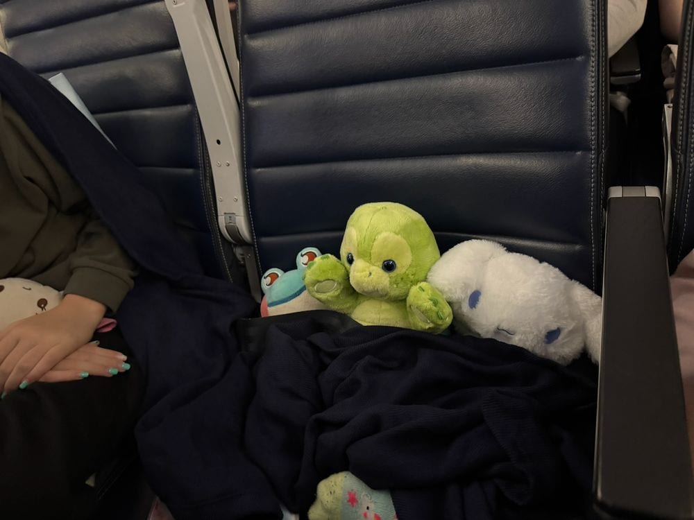Three stuffed animals, including a green turtle and a white Cinnamoroll plush, sit bundled in a dark blanket on an airplane seat next to a child's arm with teal nail polish.
