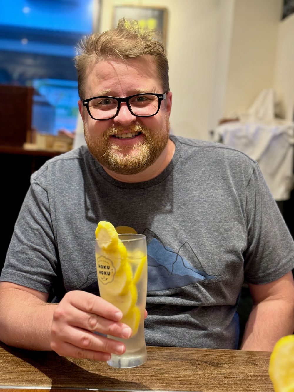 A man smiles while holding a tall glass of lemon sour drink garnished with sliced lemons at a restaurant table.