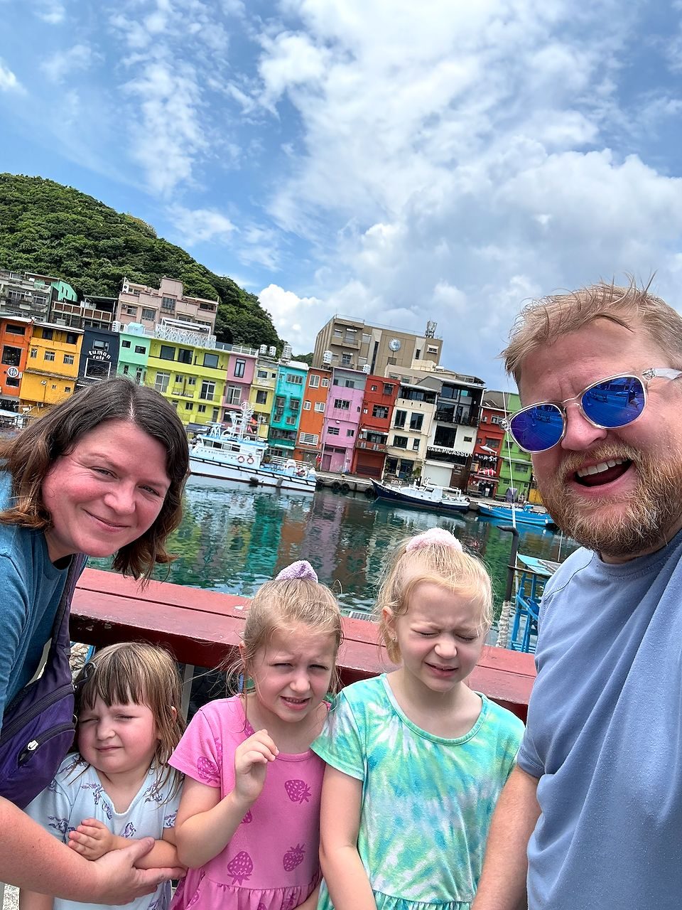 A family selfie at Zhengbin Fishing Harbor with a woman and man flanking three young girls, all squinting in the sunlight, with a row of brightly painted multi-colored buildings reflected in the harbor water behind them.