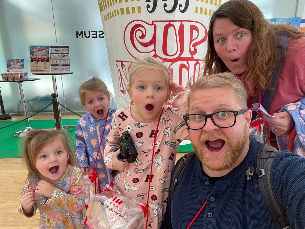 Adam takes a wide-angle selfie with Lindsay and their three daughters in front of a giant Cup Noodles display at the Cup Noodles Museum in Yokohama, all five making exaggerated open-mouthed excited faces while holding souvenir bags.