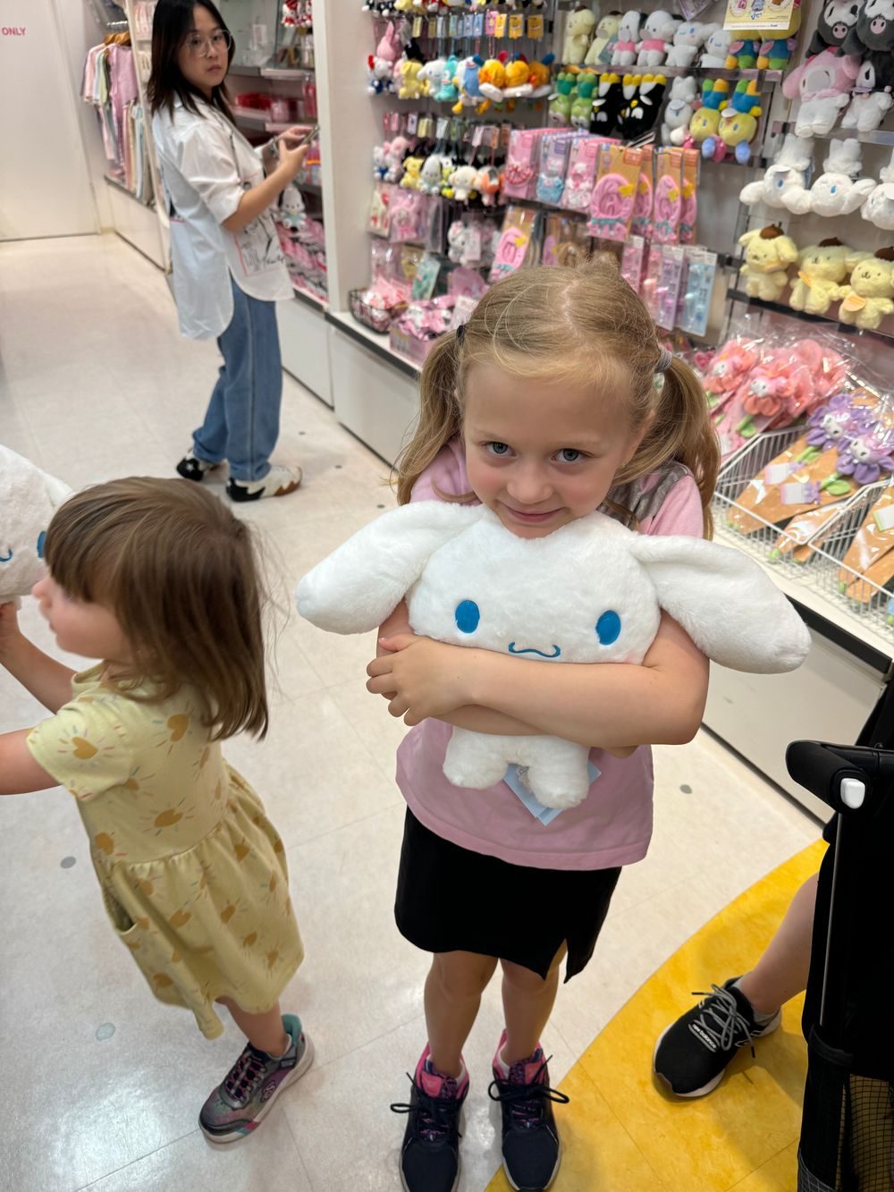 A young girl hugs a large white Cinnamoroll plush toy inside a Sanrio character store, with shelves of merchandise and another child visible behind her.