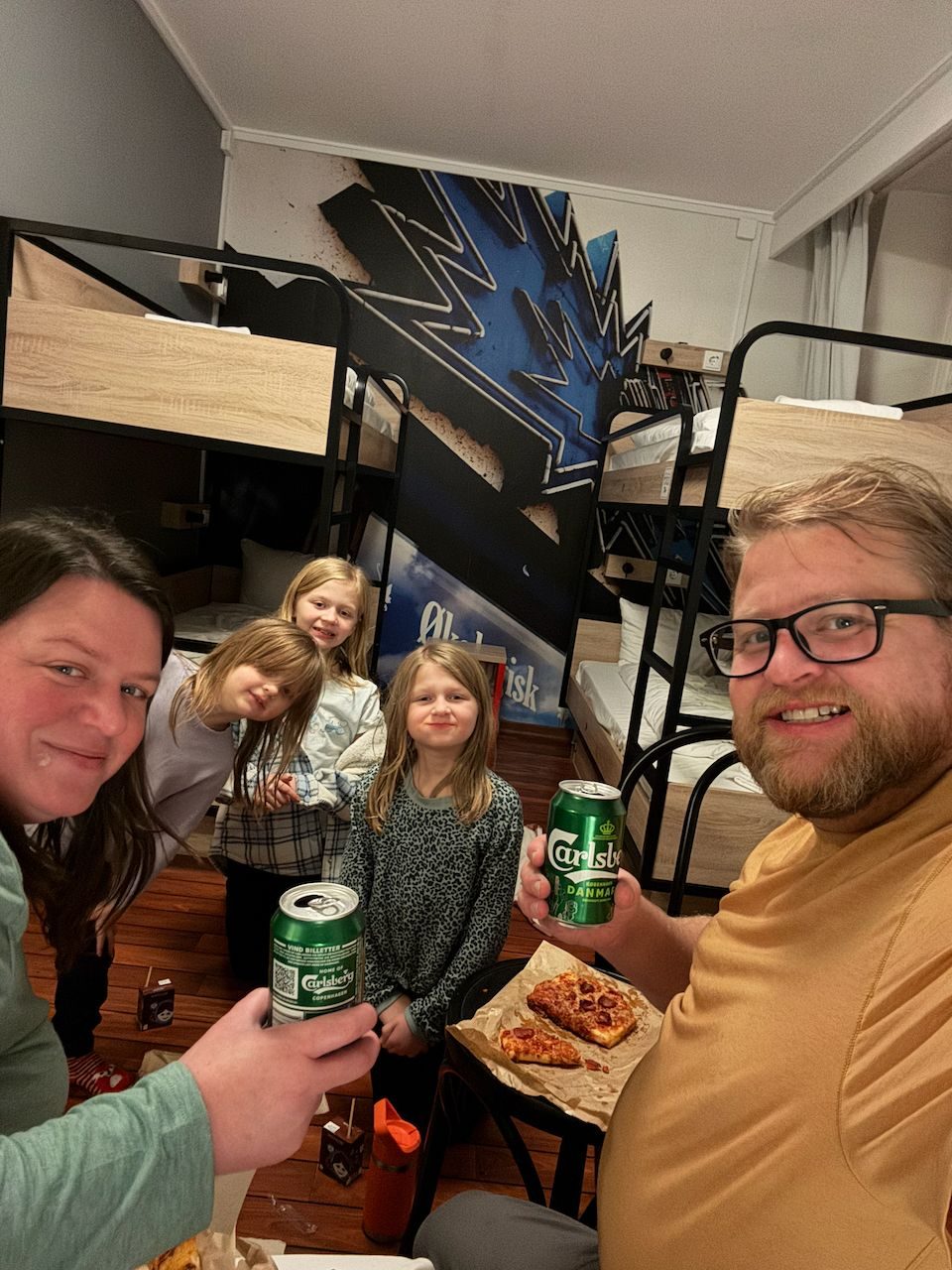 A family of five in a Copenhagen hostel room with bunk beds and graffiti murals, pizza on the table and a Carlsberg in hand
