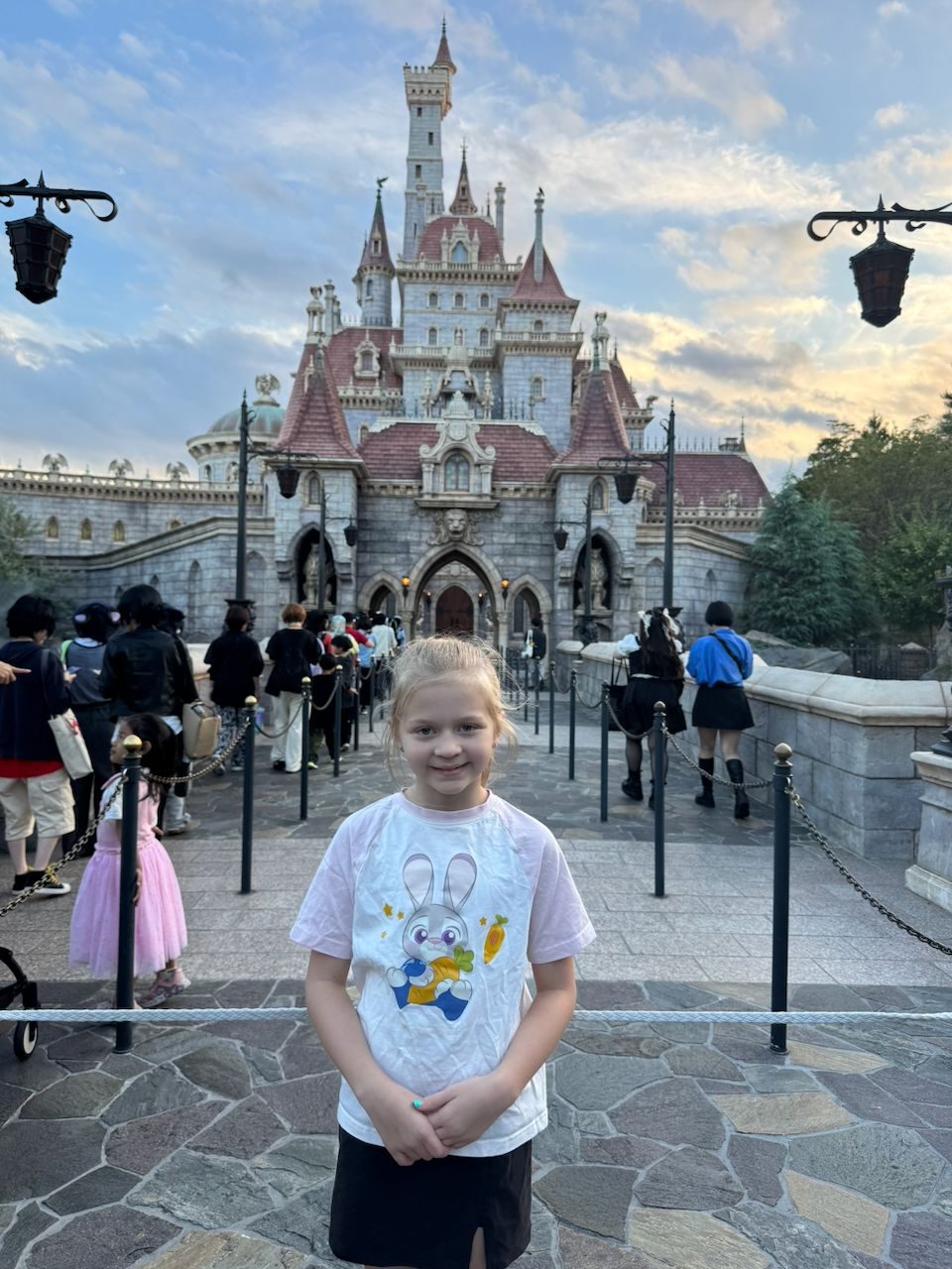 A young blonde girl stands smiling in front of the ornate Beauty and the Beast castle at Tokyo DisneySea, with a crowd of park guests queuing behind her and a partly cloudy sky overhead.
