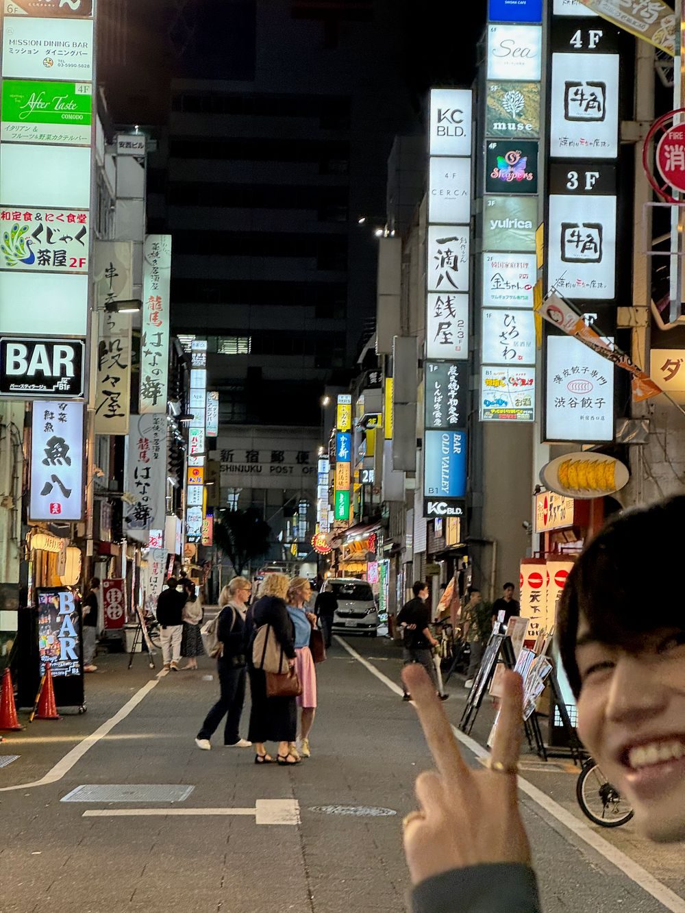 A crowded Kabukicho street at night in Tokyo, lit by stacked neon signs, with pedestrians crossing and a young man flashing a peace sign toward the camera.