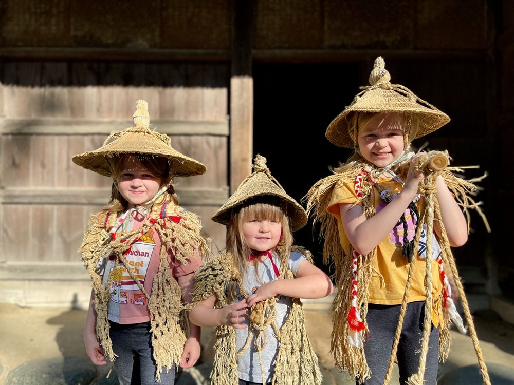 Three young girls wearing traditional Japanese straw hats and woven straw capes stand in front of a dark wooden barn, squinting in bright autumn sunlight, one holding a bundle of rope decorations.