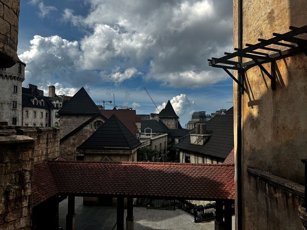 A view across the rooftops of the French village attraction at Ba Na Hills, with dark pointed rooflines and stone buildings framed against a blue sky with large white clouds, and construction cranes visible in the background.