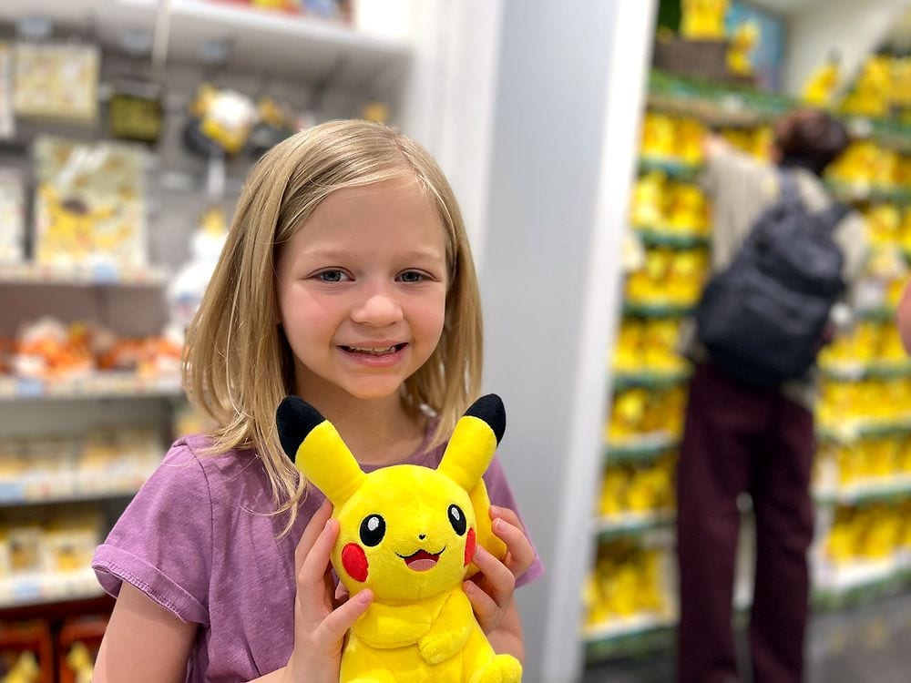 A young blonde girl in a pink shirt smiles and holds up a Pikachu plush toy inside a Pokémon Center store, with shelves of yellow merchandise visible behind her.