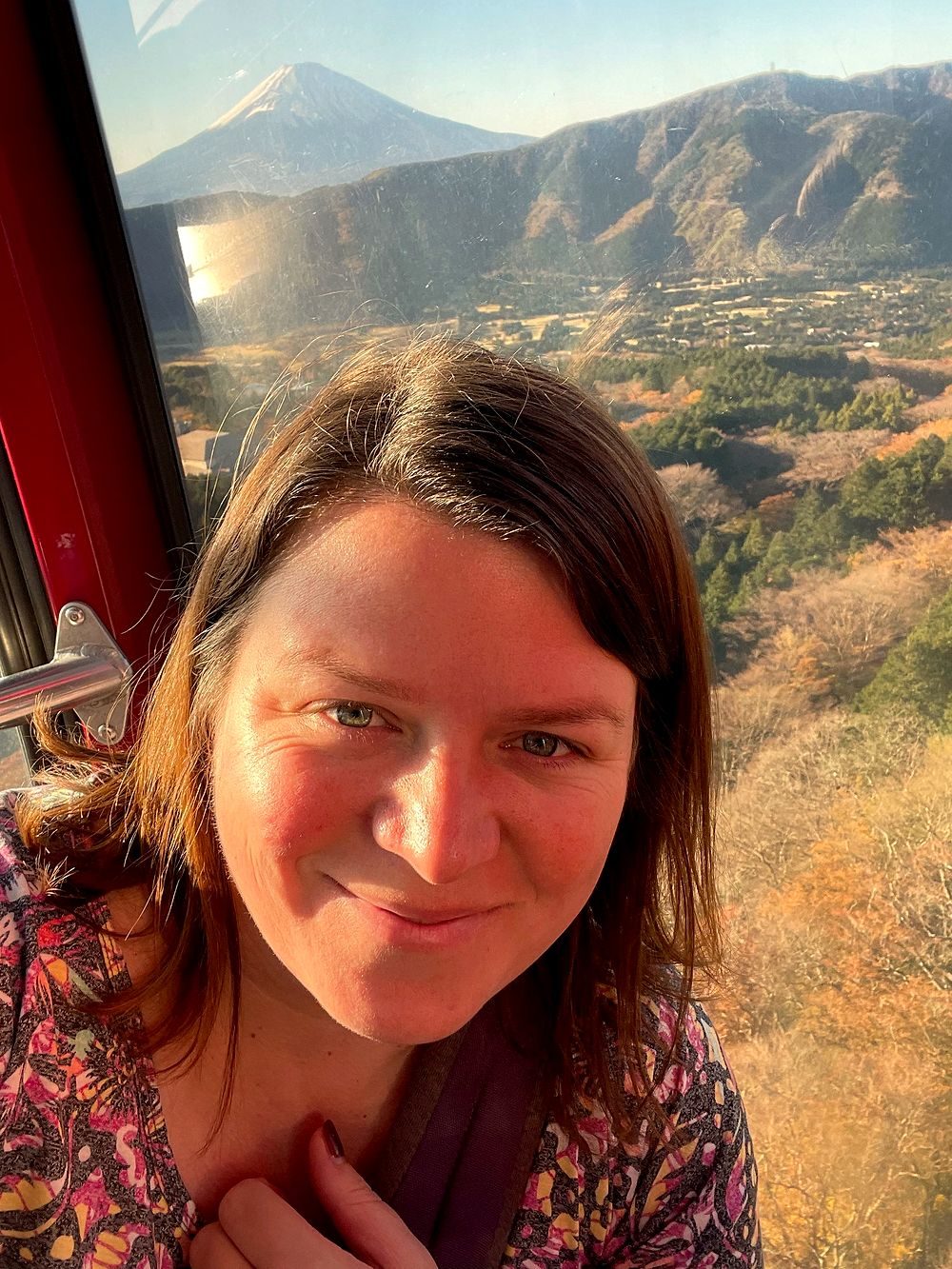 A woman takes a selfie inside a Hakone Ropeway gondola with Mount Fuji and the surrounding valley visible through the window behind her.