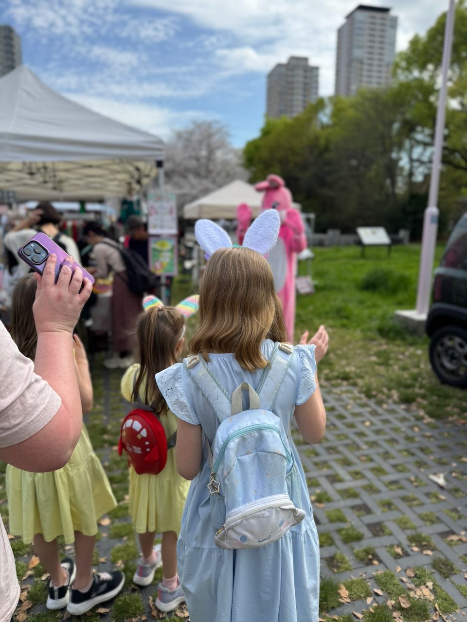 Two girls wearing bunny ears walk toward a person in a pink Easter bunny costume at an outdoor market in Osaka, with cherry blossom trees and market stalls visible in the background.