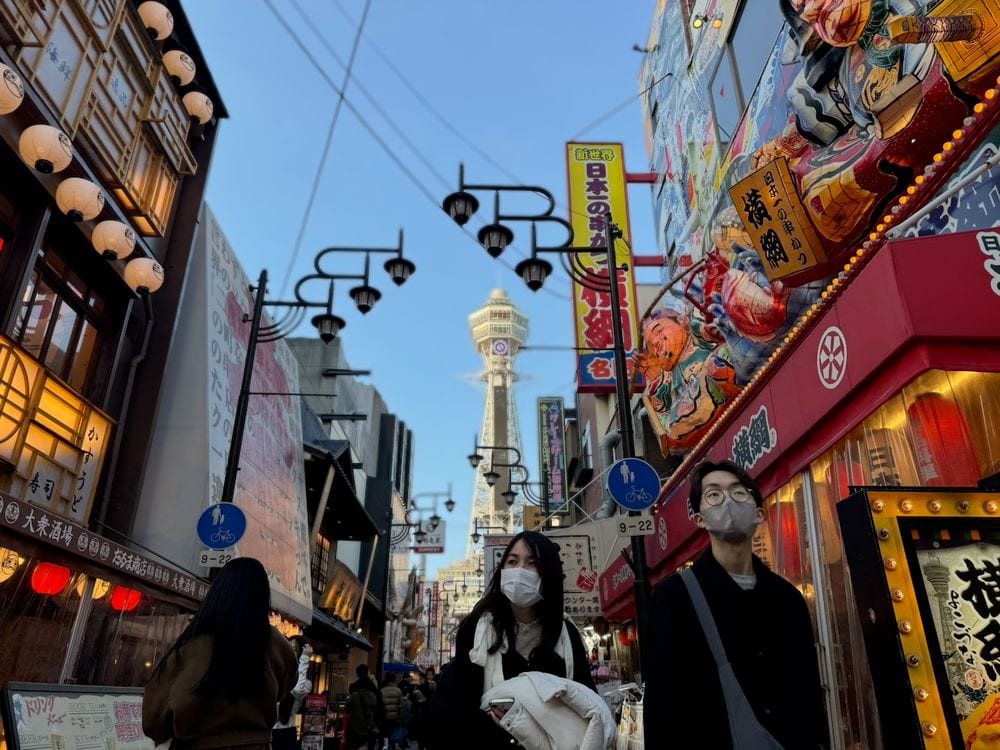 A busy Shinsekai shopping street in Osaka lined with lanterns and colorful restaurant signs, with Tsutenkaku tower at the far end under a clear blue sky