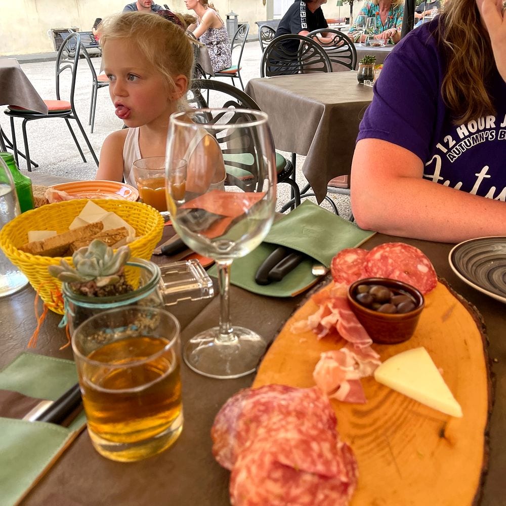 A young girl sticks her tongue out at an outdoor restaurant table in Tuscany, with a wooden charcuterie board of salami, prosciutto, olives, and cheese in the foreground