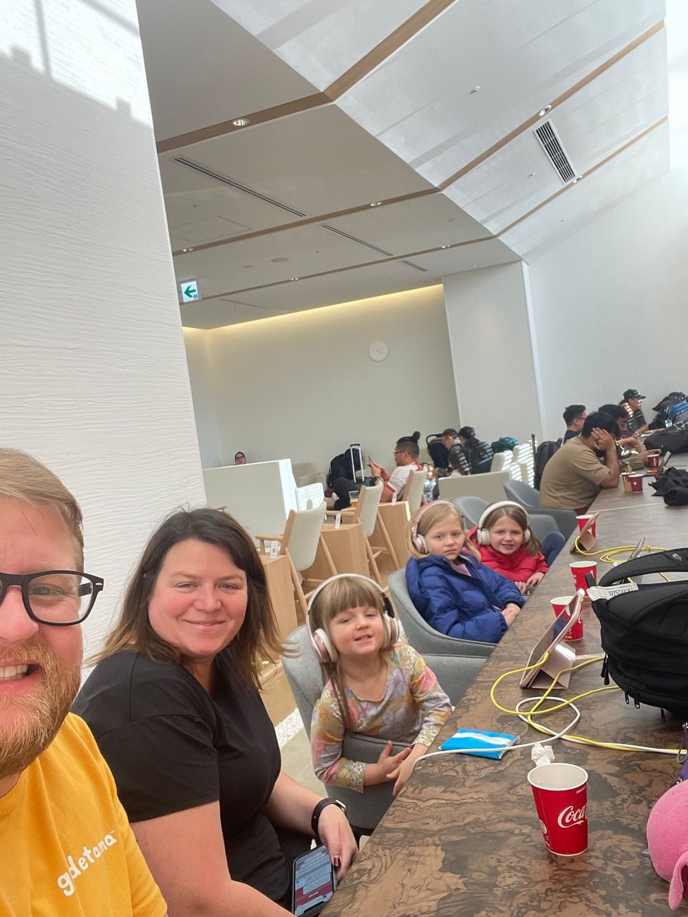 A family sits in a quiet airport lounge at Narita Airport, two young girls wearing headphones at a long wooden table.