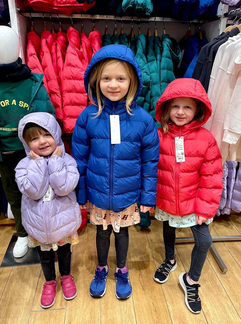 Three young girls try on brightly colored puffer jackets with price tags still attached inside a clothing store, surrounded by racks of winter coats.