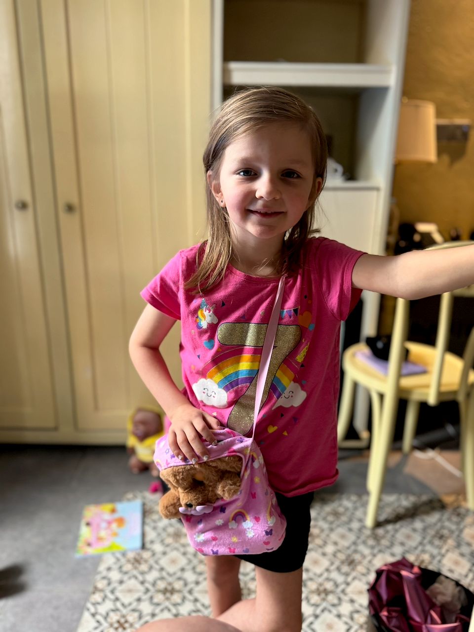 A young girl in a pink rainbow birthday shirt stands smiling at the camera, holding a small pink purse with a stuffed animal inside.