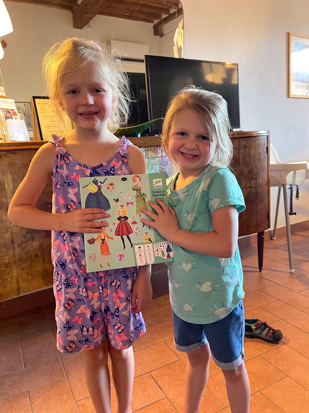 Two young blonde girls stand in a Tuscan farmhouse interior, smiling and holding up Djeco sticker sets and paper doll gifts left by their Airbnb host