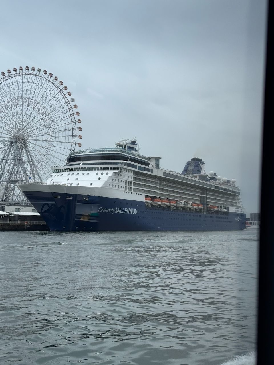 The Celebrity Millennium cruise ship docked at Osaka's Sakurajima port, with the large Tempozan Ferris Wheel visible behind it and grey water in the foreground, shot through a window.