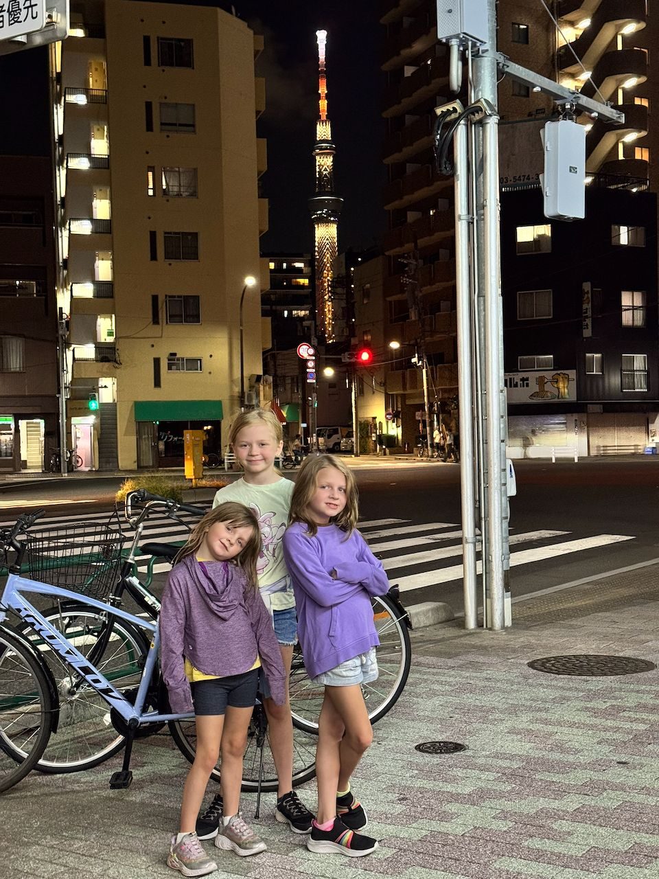 Three girls standing together on a Tokyo sidewalk at night with parked bicycles beside them and the illuminated Tokyo Skytree glowing orange in the background.