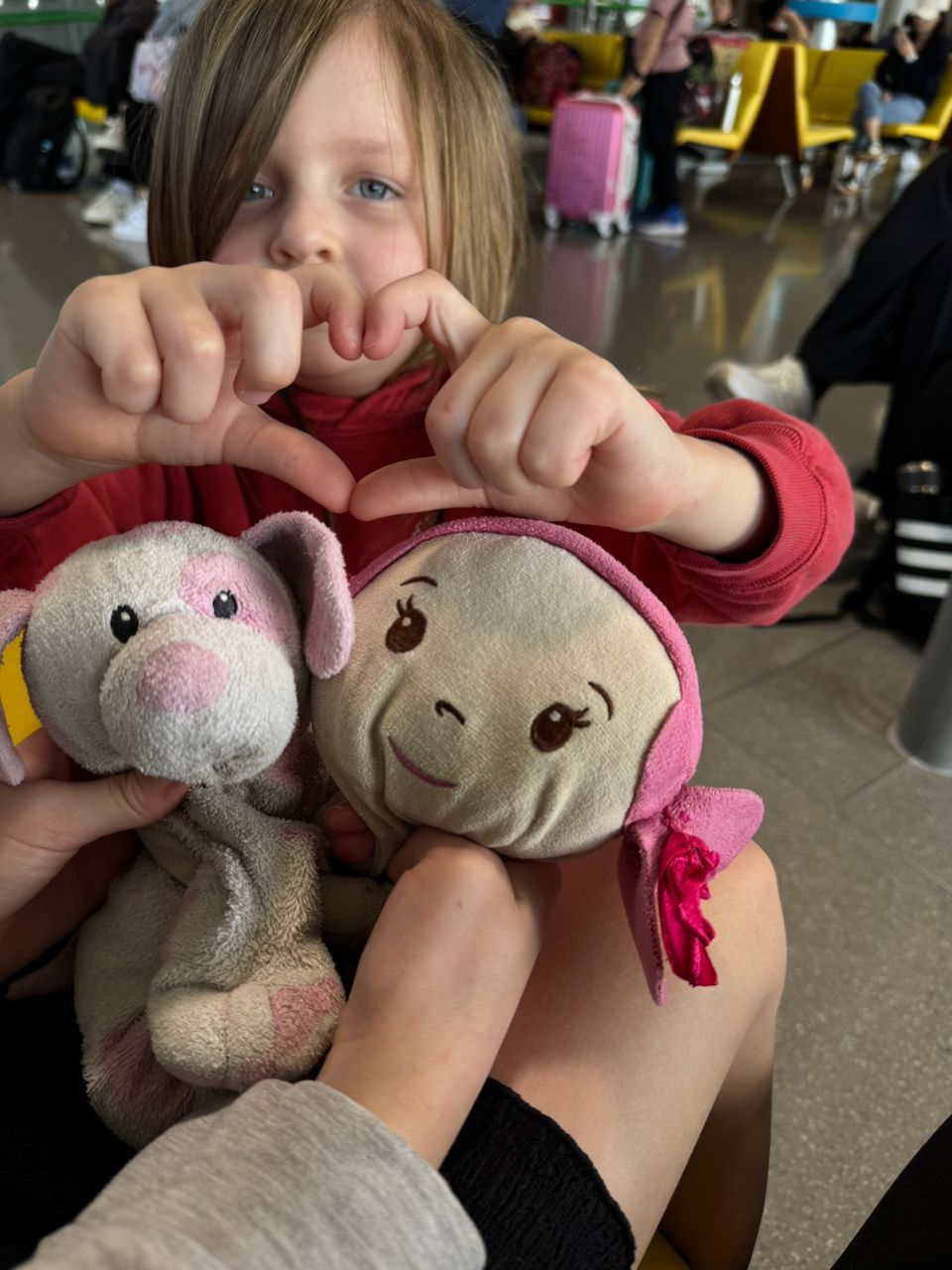 A young girl sits in an airport terminal holding two small stuffed animals and making a heart shape with her fingers above them, with luggage and other travelers visible in the background.