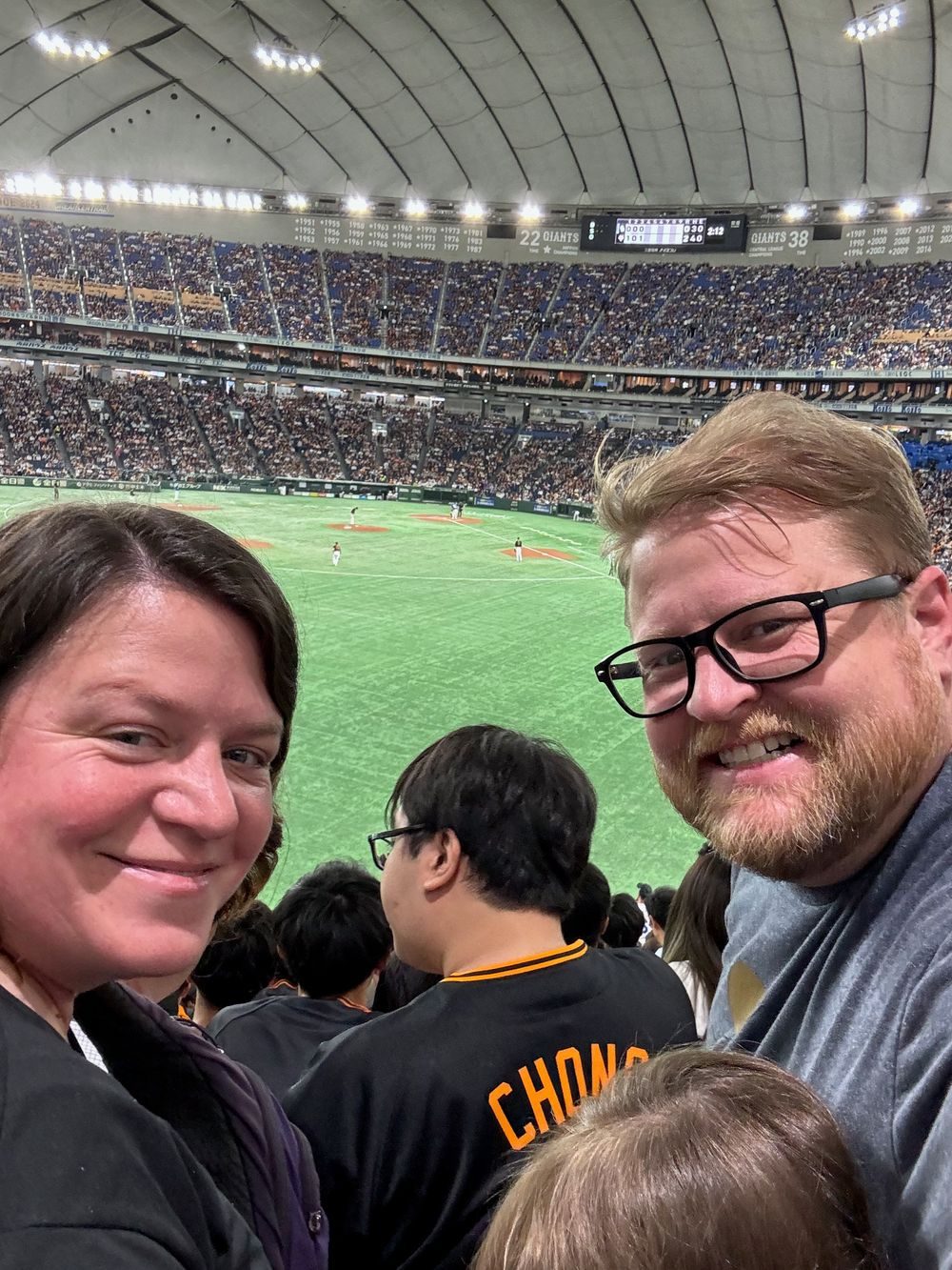 A man and woman smile in stadium seats with a baseball field visible below them at Tokyo Dome.