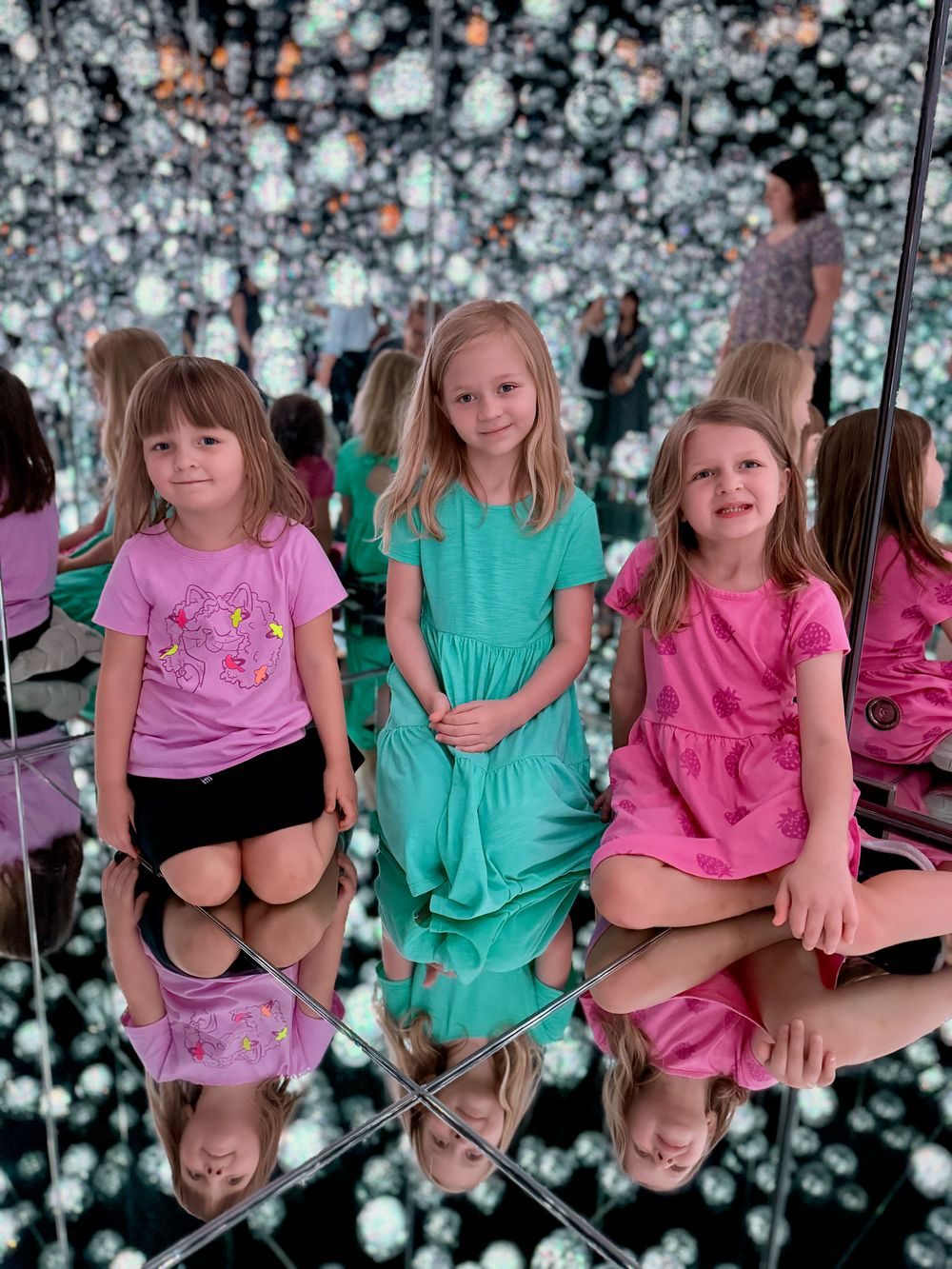 Lily, Cora, and Harper sitting side by side on a mirrored floor inside a TeamLab installation, surrounded by infinite reflections of glowing white orbs