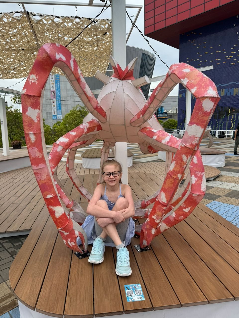 A young girl with glasses sits cross-legged and smiling inside the claws of a large decorative red crab sculpture on a wooden deck at the Osaka waterfront, with a blue tiled building visible behind her.