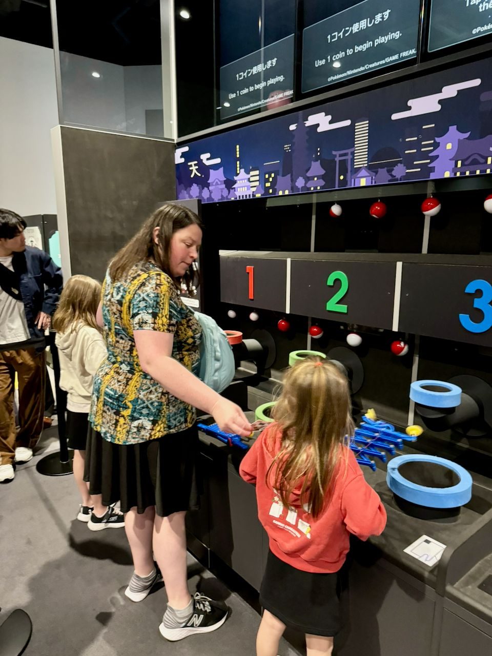 Lindsay stands at a coin-operated Pokémon arcade-style ring toss game, helping a young girl in a red shirt aim at numbered targets decorated with Pokéballs, inside a dark exhibition space with Japanese and English signage overhead.