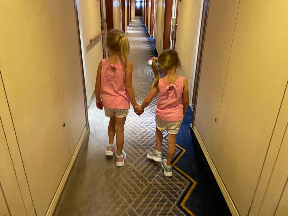 Two young girls walking hand in hand down a long ship corridor toward the kids zone on the Queen Mary 2