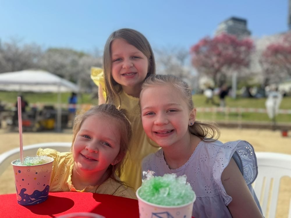 Three of our girls with shaved ice at an outdoor table under cherry blossoms at Ogimachi Park, Osaka, Japan, April 2026