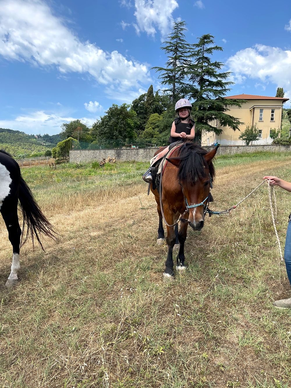 A young girl wearing a pink riding helmet sits atop a brown horse being led by a guide, with Tuscan hills, trees, and a farmhouse in the background