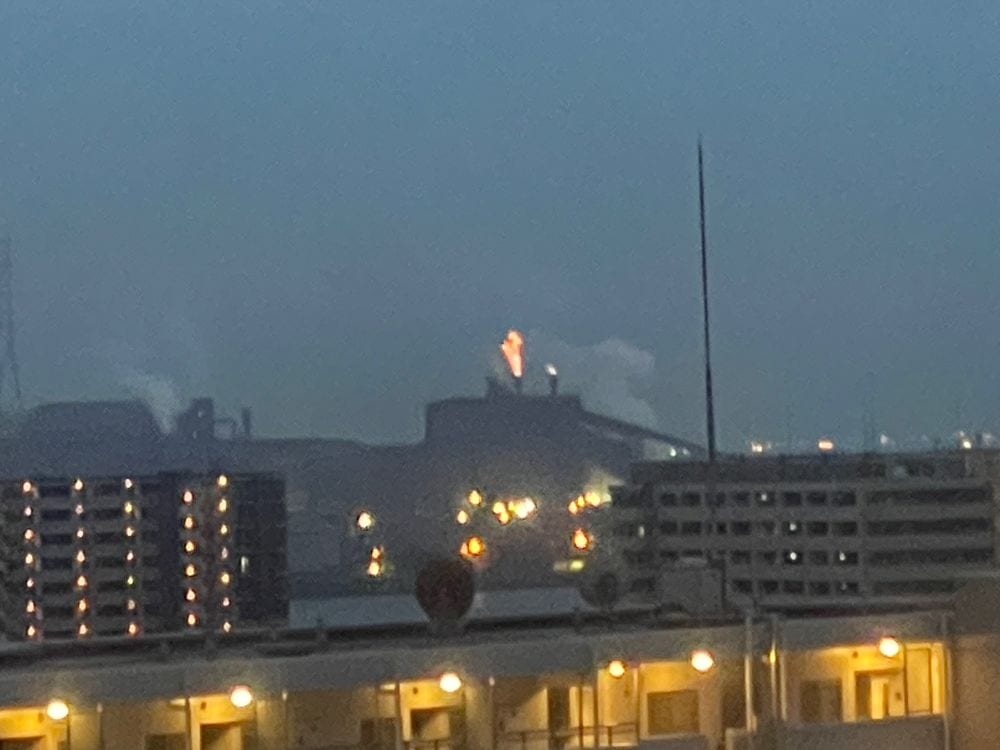 A nighttime industrial skyline in Chiba, with a bright glow or flame rising from one building among illuminated factory structures.