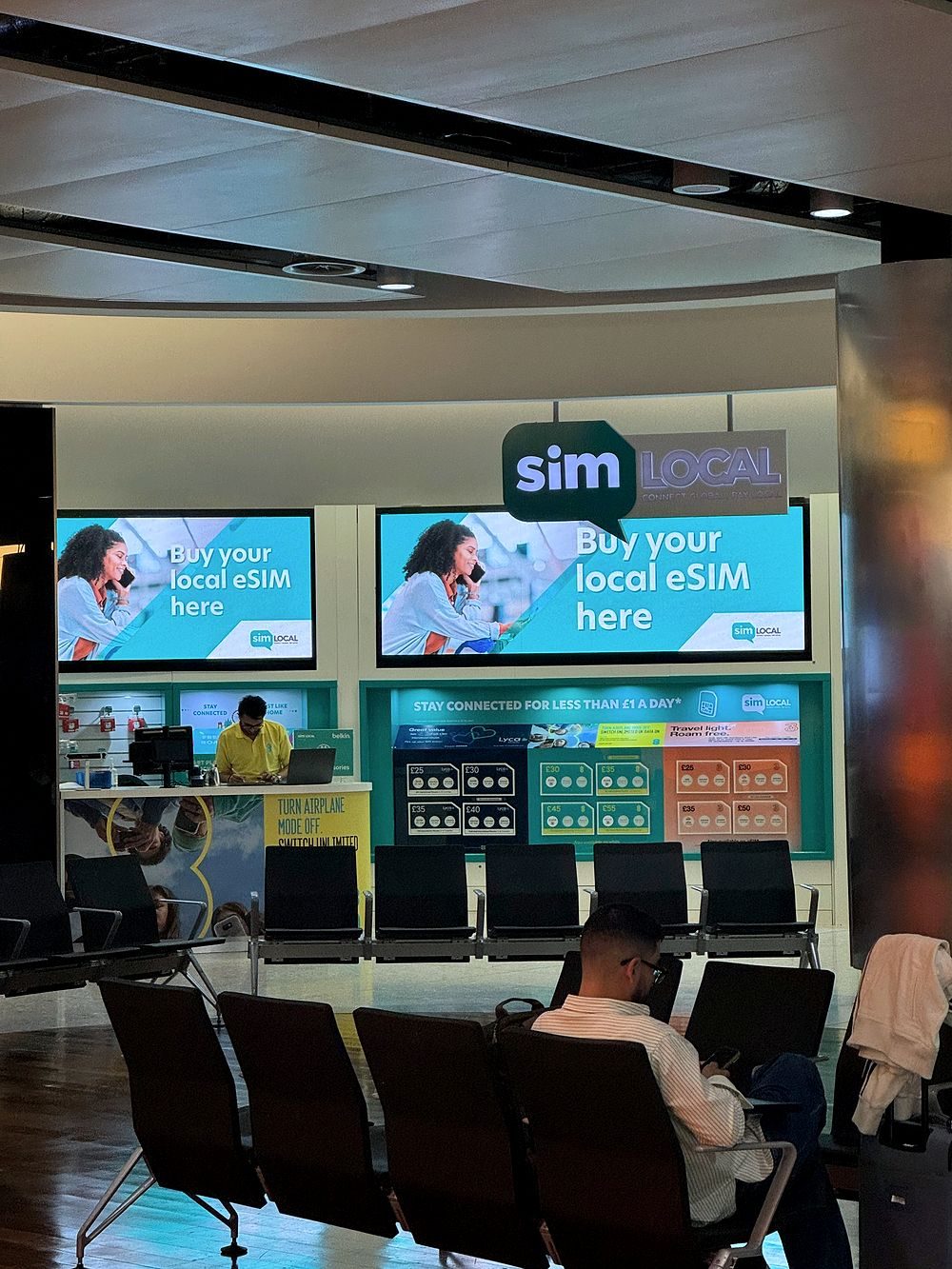 A SimLocal eSIM retail kiosk inside Heathrow Airport with screens reading "Buy your local eSIM here," a staffed counter, and SIM card display racks