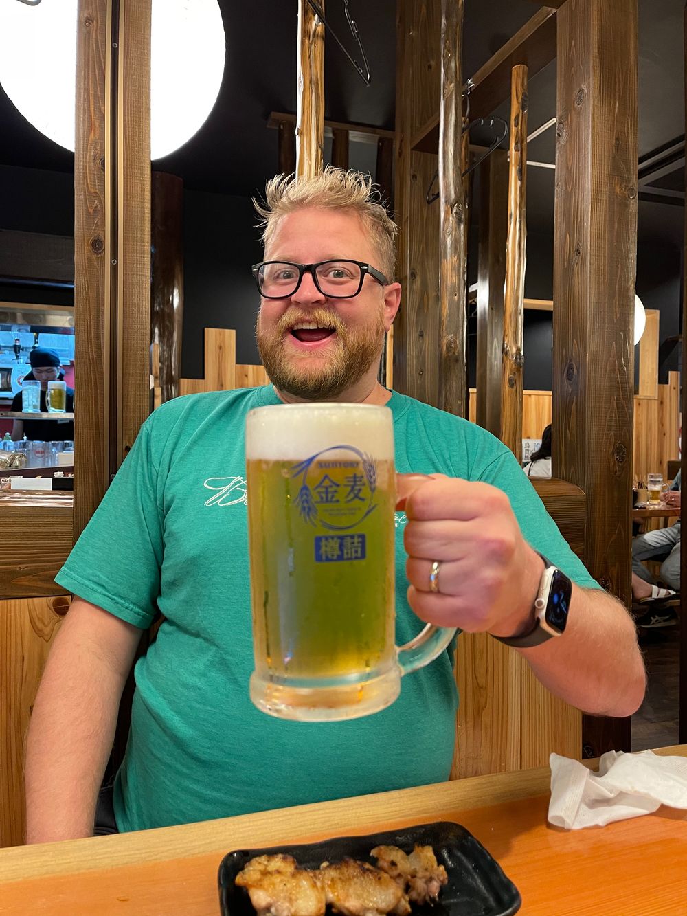 Adam holds up a large frosted mug of Suntory Kinmugi draft beer toward the camera with a wide grin, sitting at a wooden bar with a tray of yakitori visible in the foreground.