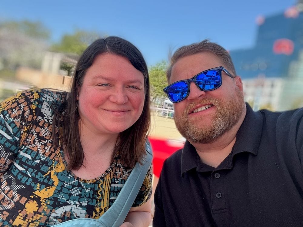Adam and Lindsay take a selfie outdoors on a sunny day in Osaka, with blue sky and a blurred building visible behind them.