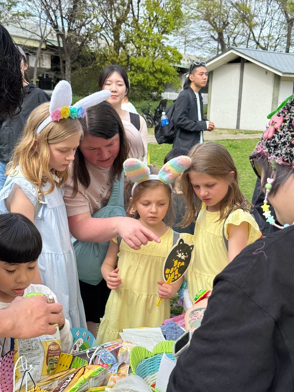 A woman and three young girls wearing bunny ear headbands crowd around an outdoor activity table at what appears to be an Easter event in a park in Osaka, Japan, with the girls examining items on the table.