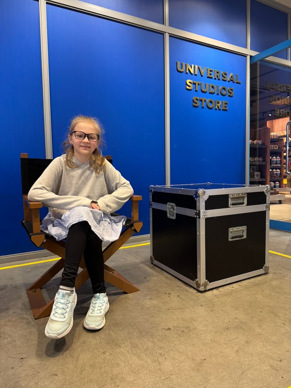 A young girl with glasses sits confidently in a wooden director's chair in front of the blue Universal Studios Store entrance at Universal Studios Japan.