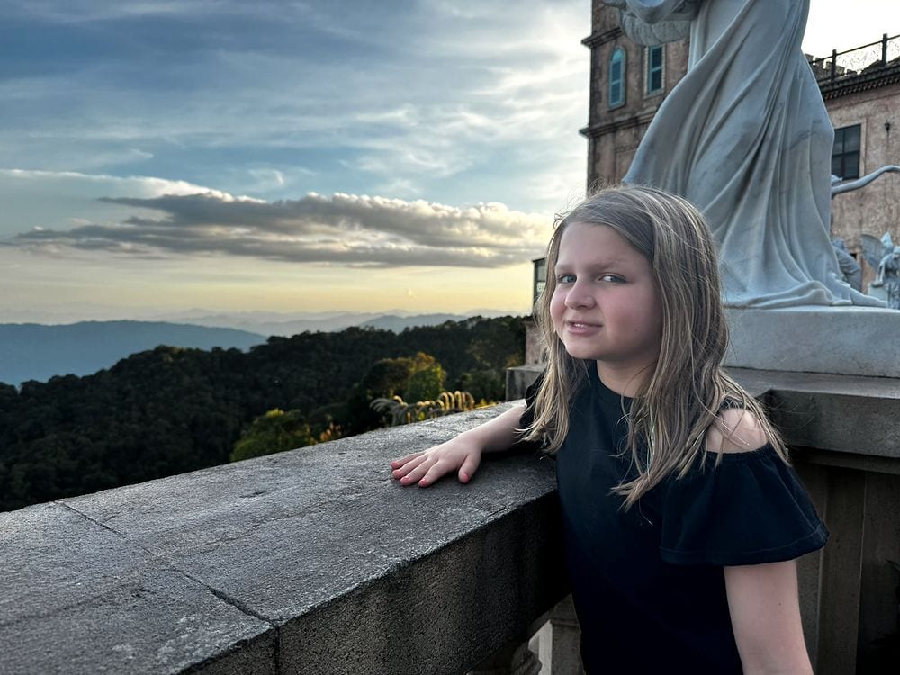 A girl leans against a stone balcony railing at Ba Na Hills, looking out toward a forested mountain ridgeline under a golden hour sky, with a large white statue and pink castle building visible behind her.