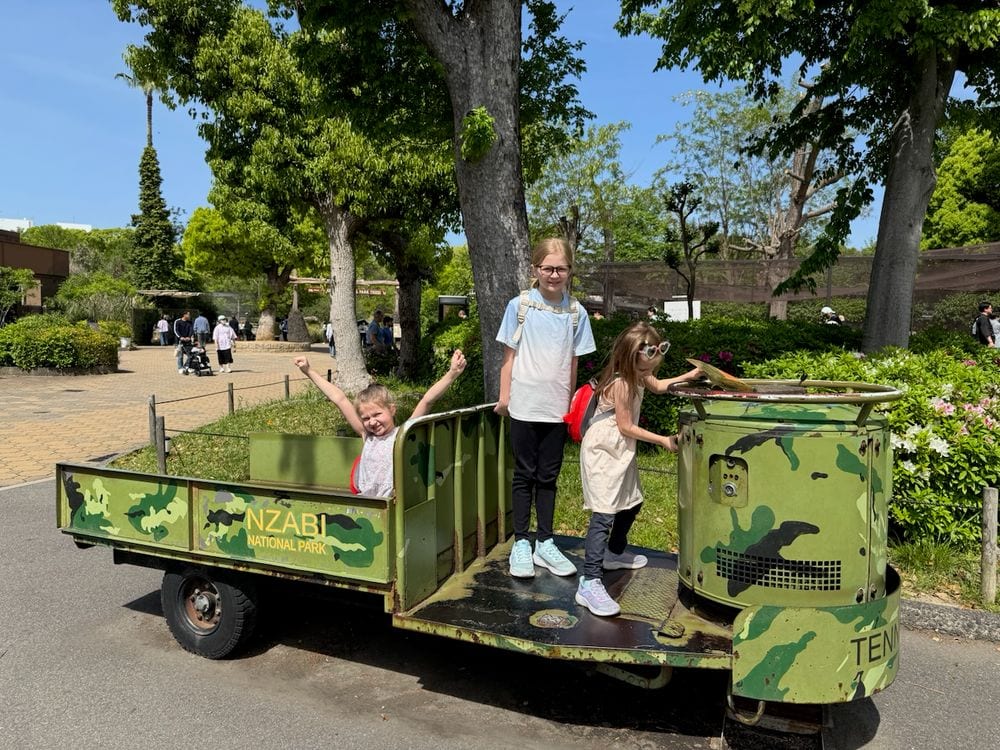 Three girls pose on a camouflage-painted safari-style vehicle marked Nzabi National Park at an outdoor zoo on a sunny spring day.