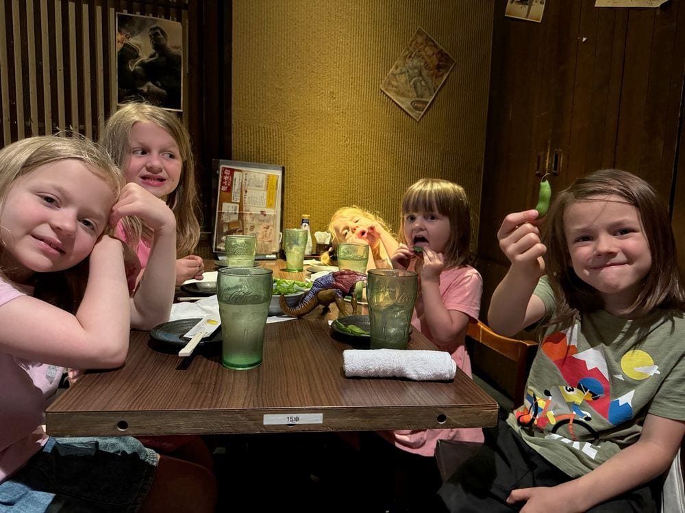 Five girls sit around a wooden izakaya table in Tokyo, several holding bright green melon soda drinks, with plates of food and edamame on the table.