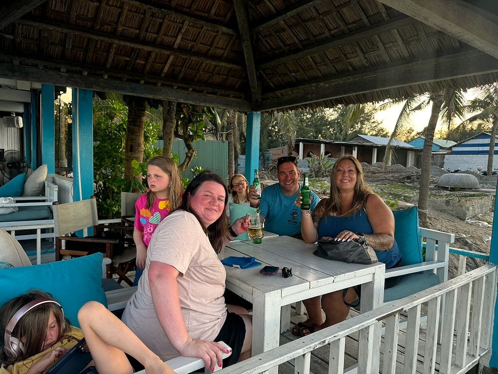 Adults and children around a table under a thatched beach bar pavilion in Hoi An at sunset, with beer bottles on the table and a girl in the foreground watching an iPad