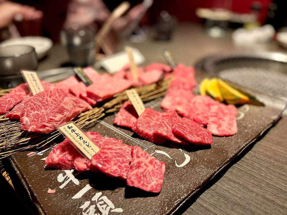 Seared wagyu beef skewers and a grilled wagyu burger patty on a teppanyaki grill, with a water glass and mesh strainer visible on the counter.