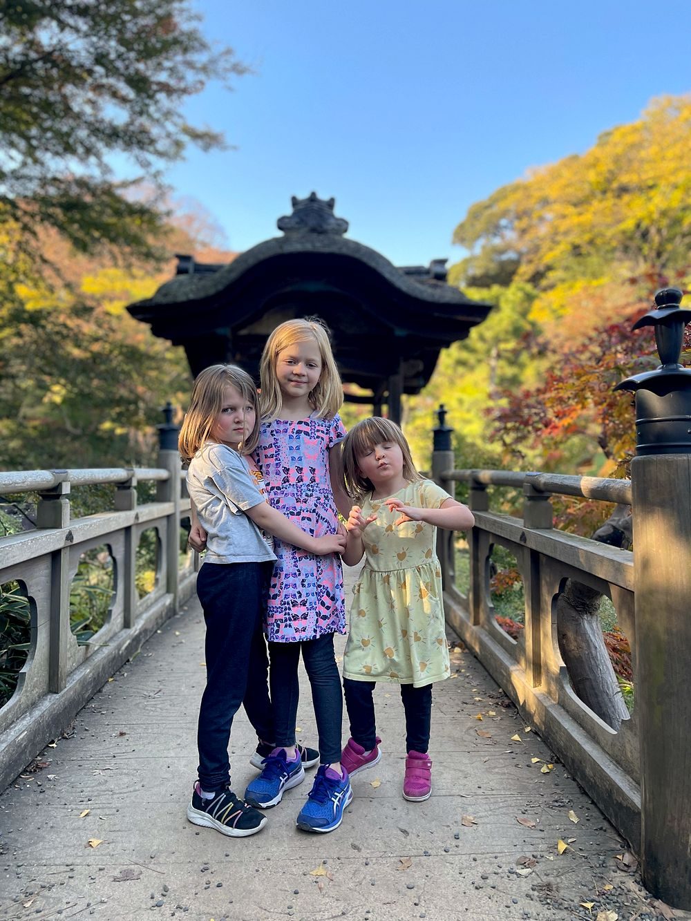 Three young girls stand together on a wooden bridge in front of a traditional pavilion at a Japanese garden, autumn foliage visible around them under a clear blue sky.