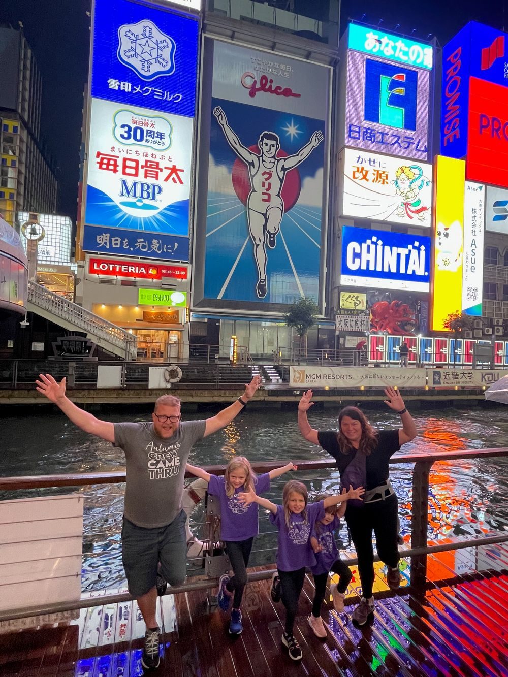 A family of five raises their arms on a boat deck in front of the illuminated Glico Man billboard at Dotonbori, Osaka, at night.