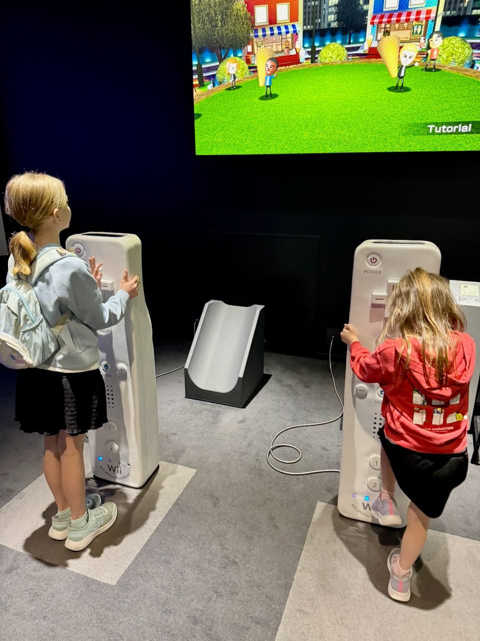 Two girls stand facing a large screen showing a Wii tutorial, each holding oversized life-size Wii remote props in a darkened museum exhibit room.
