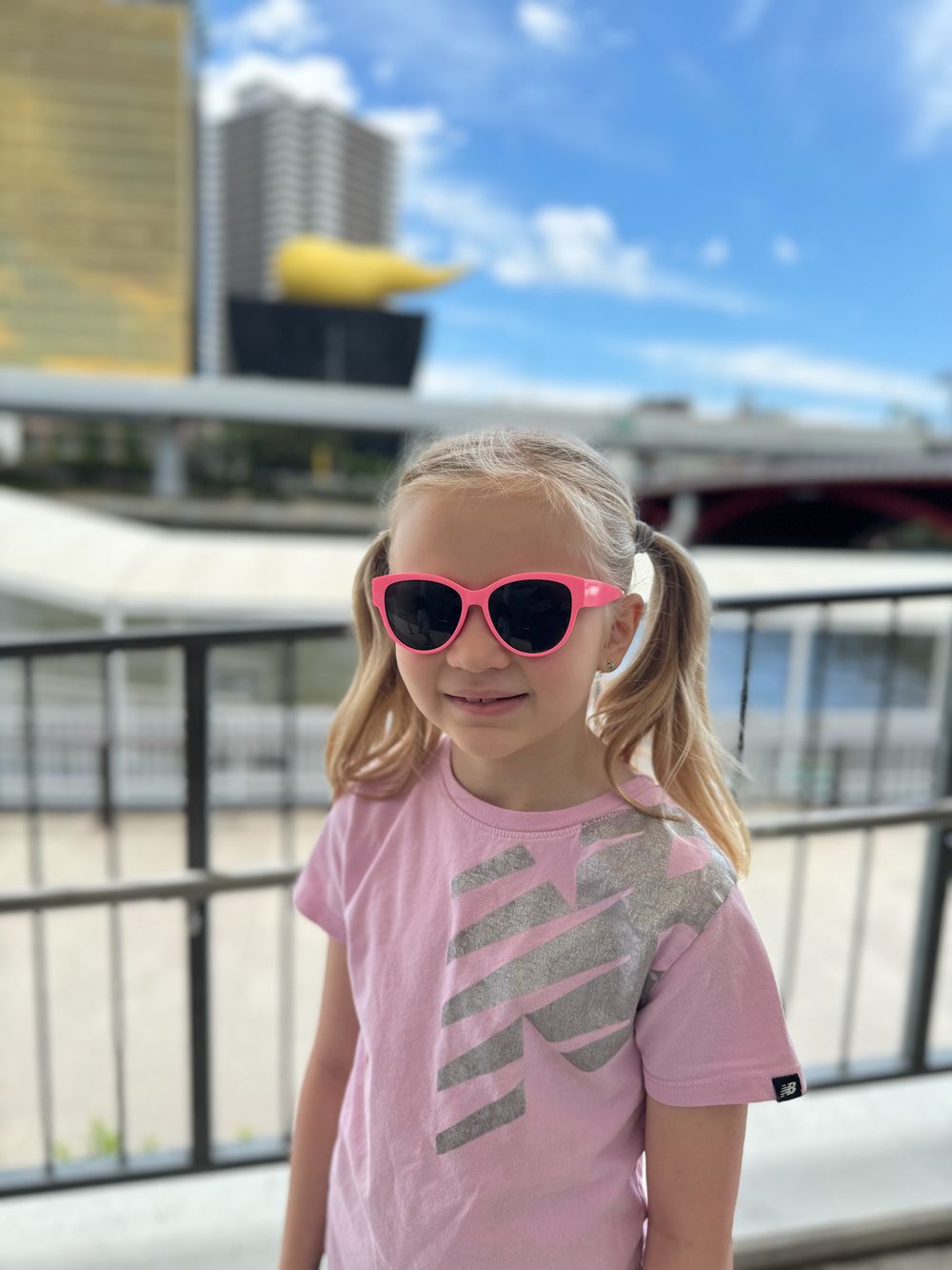 A young girl wearing pink sunglasses stands along the Sumida River in Asakusa, with the golden Flamme d'Or sculpture visible on top of the Asahi building behind her.