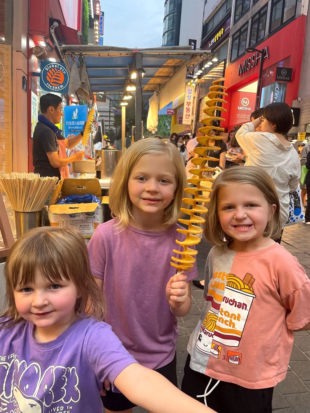 Three young girls on a busy Myeongdong street at dusk, one holding a long spiral potato skewer, with lit street food stalls behind them